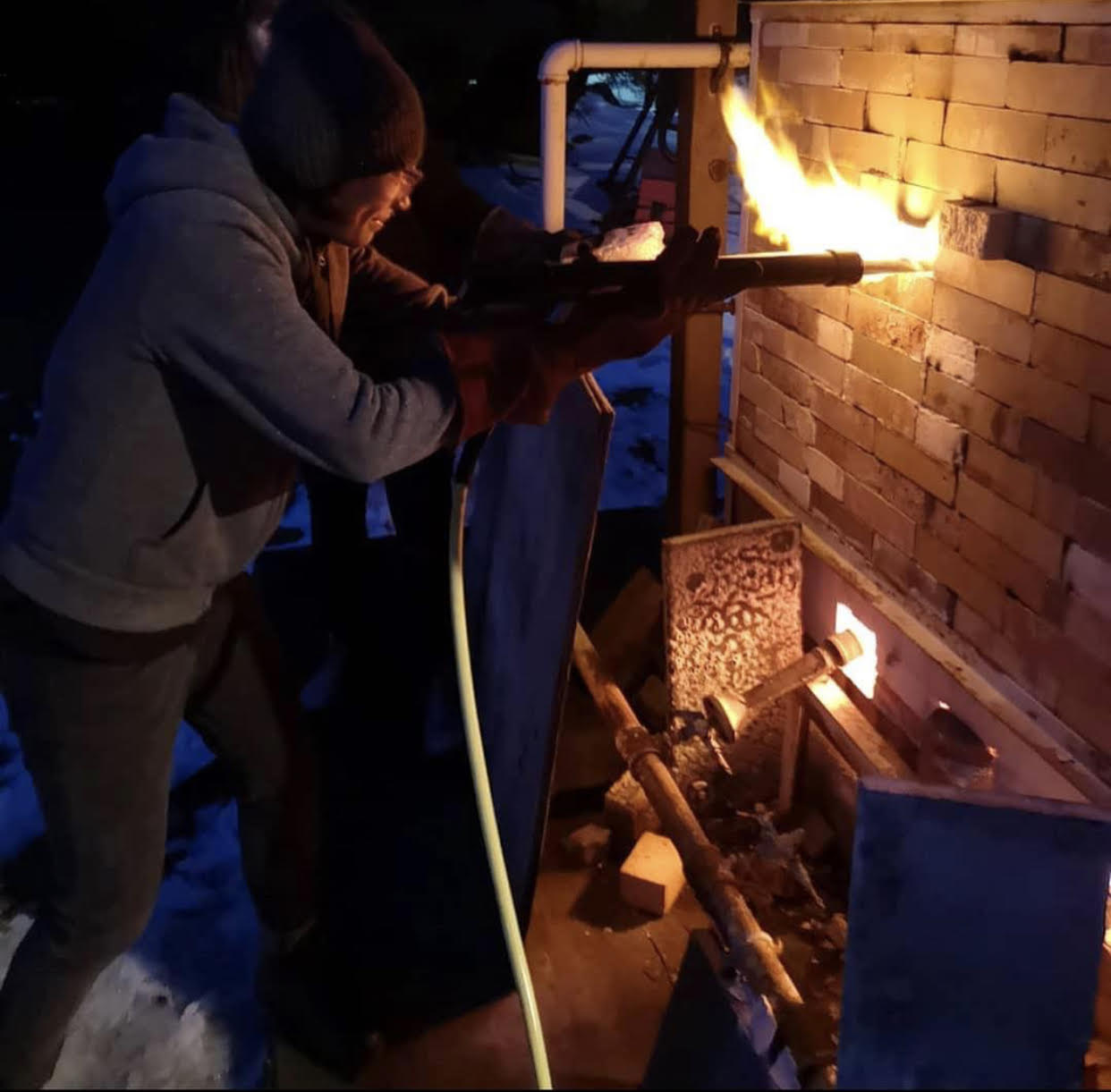Artist using handmade soda gun to shoot dry soda ash through an open hole into the kiln where flames are shooting out from.